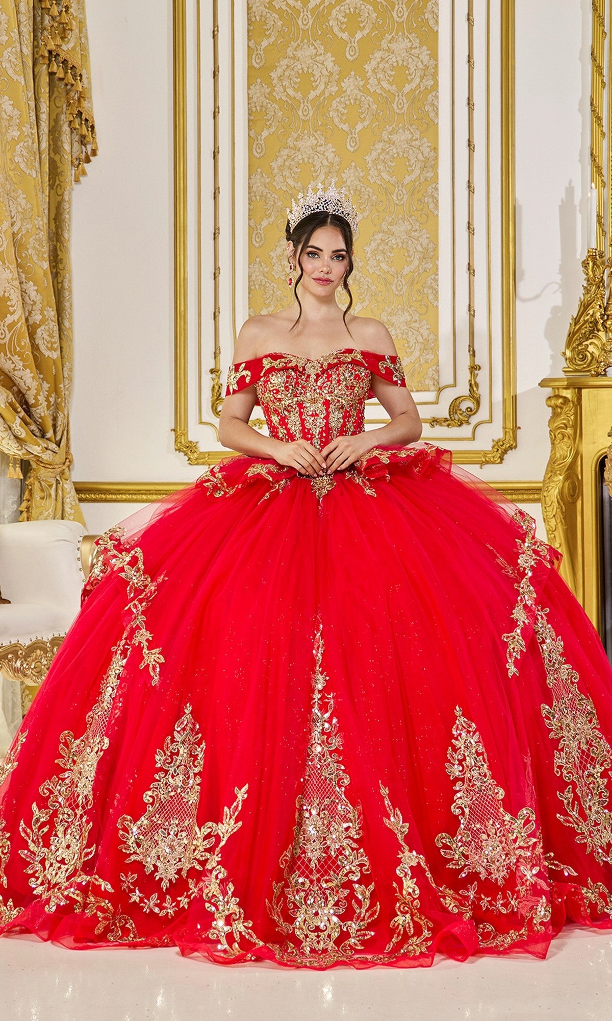 A woman in the Quinceanera Dress 5086 By Dancing Queen, a red gown with gold embroidery and a lace-up corset, stands in an ornate room with gold accents.